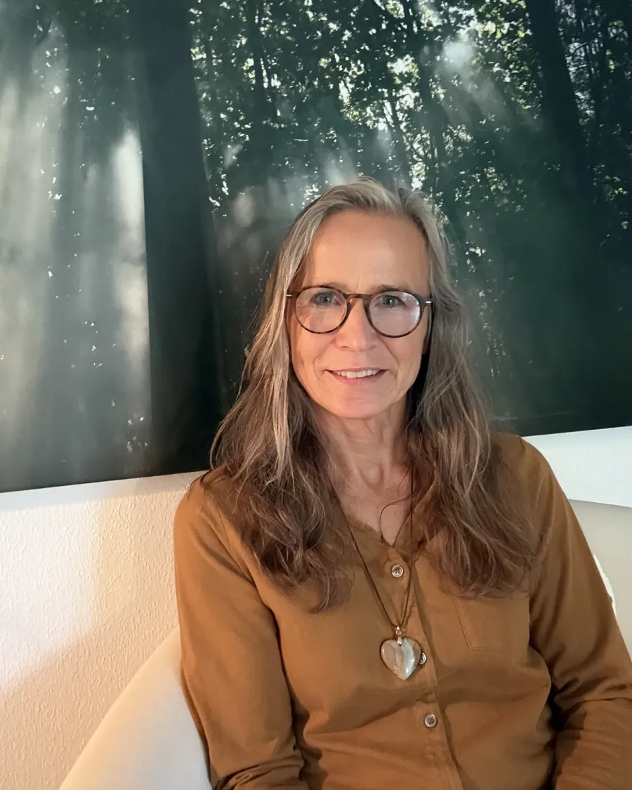 Grazyna von Lonski mit Brille im Praxisraum, hinter ihr ein großes Bild von Sonnenstrahlen im Wald