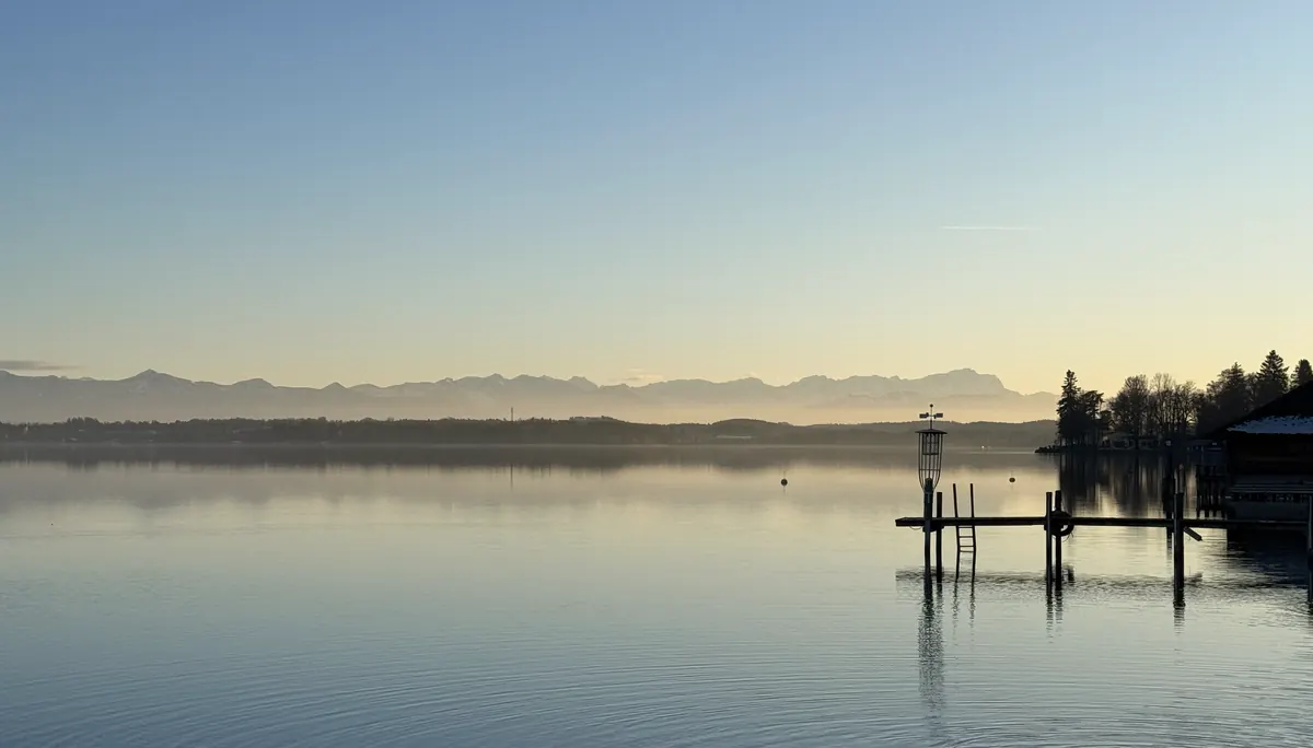 Morgenstille am Starnberger See: ein Steg im spiegelglatten Wasser, dahinter die Alpenkette im ersten Licht