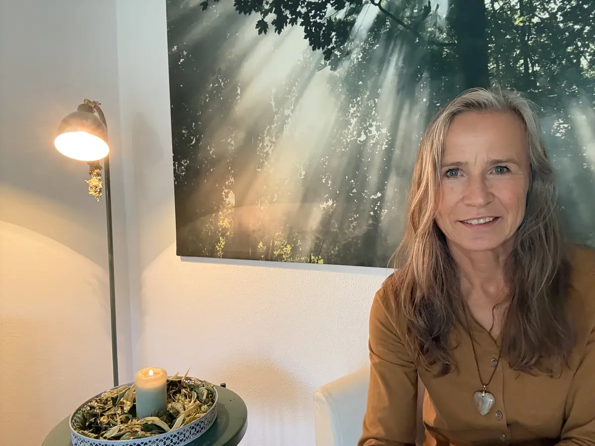 Grazyna von Lonski lächelt im Praxisraum, neben ihr Leselampe und Kerze, hinter ihr ein großes Waldbild