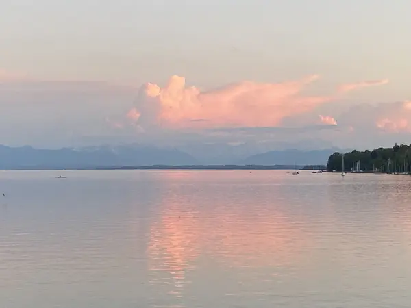 Eine rosa leuchtende Wolke spiegelt sich im glatten See, dahinter die Berge