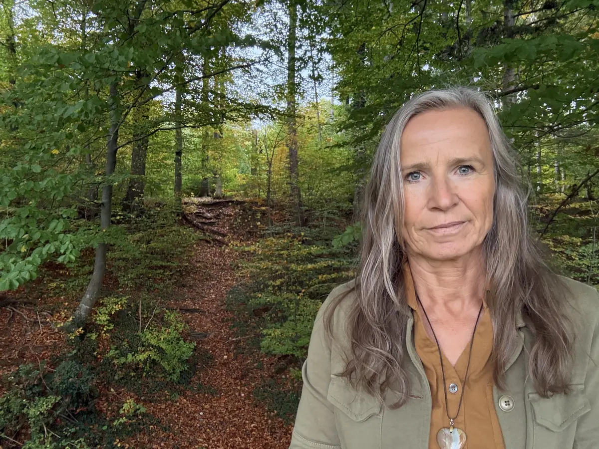Grazyna von Lonski mit ruhigem, klarem Blick im Herbstwald, neben ihr führt ein steiler Waldweg hinauf ins Licht